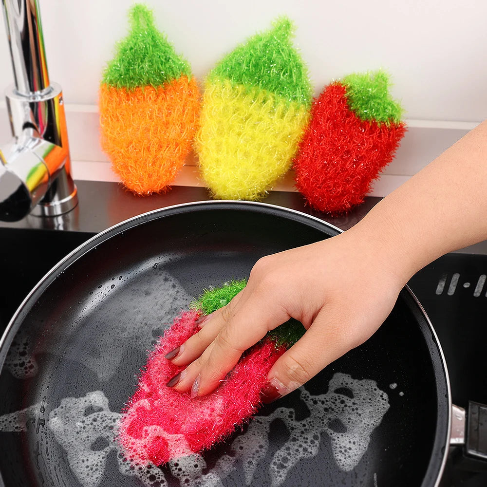A hand is using a red strawberry scrubber sponge to clean dishes in a black frying pan.