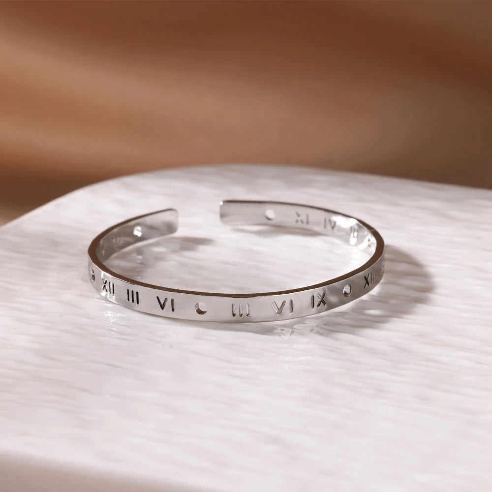 A silver-toned stainless steel cuff bracelet with Roman numerals is displayed on a white surface.