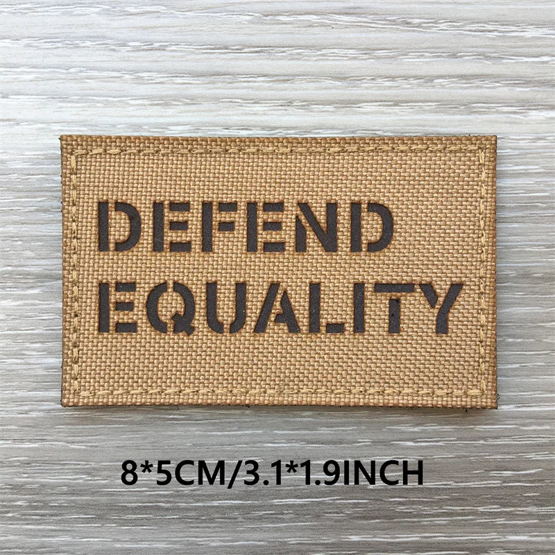 A tan woven patch with the text DEFEND EQUALITY embroidered on it, displayed on a wooden surface.