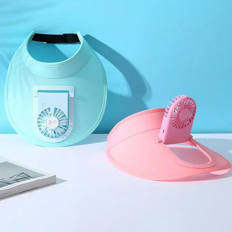 A pink cooling fan hat is displayed on a white surface next to a light blue silicone hat.