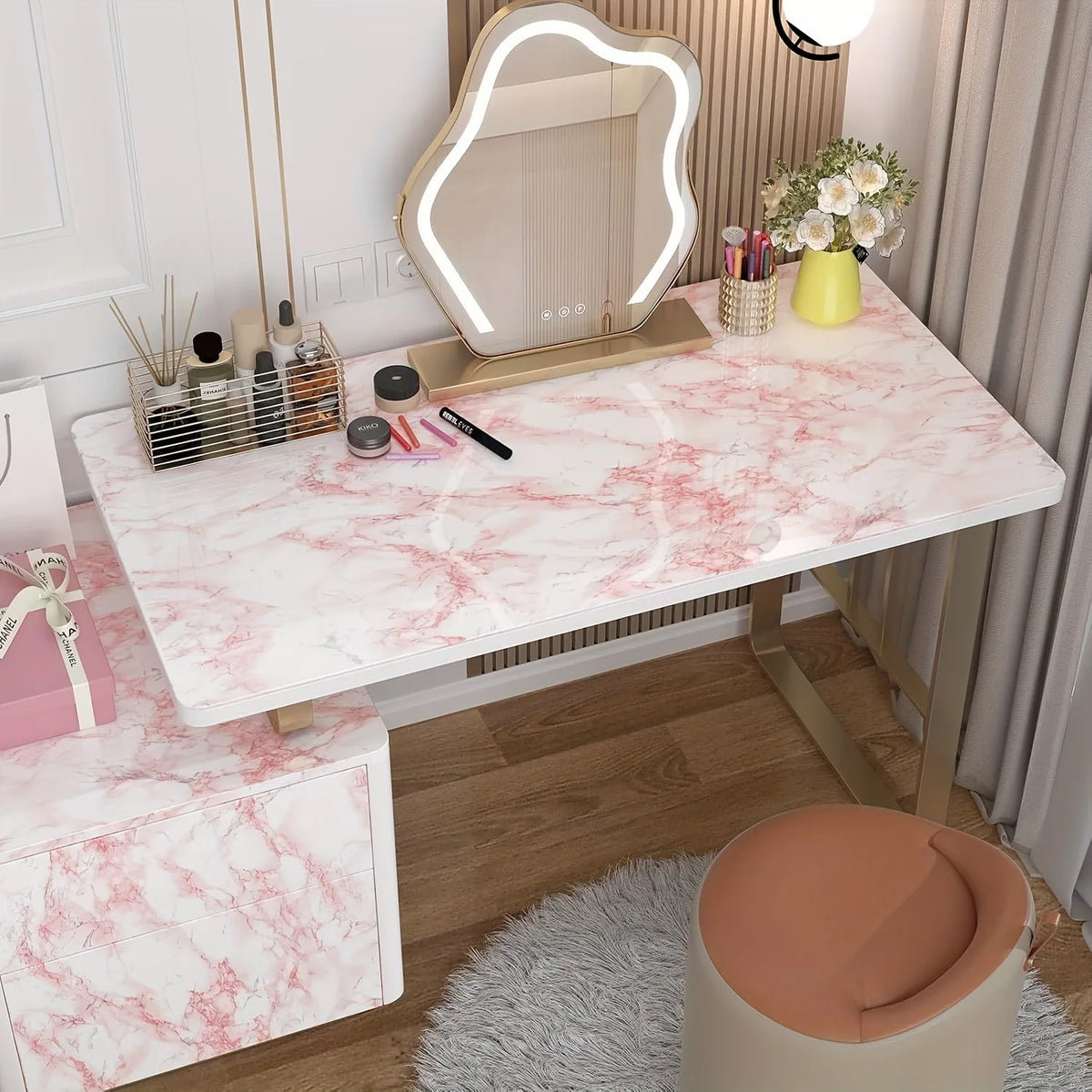 A pink marble-patterned desk surface with various cosmetic products and a lighted mirror in a bright room.