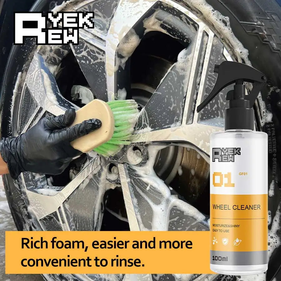 A hand is using a green brush to clean a wheel with yellow liquid foam from a white bottle of wheel cleaner.
