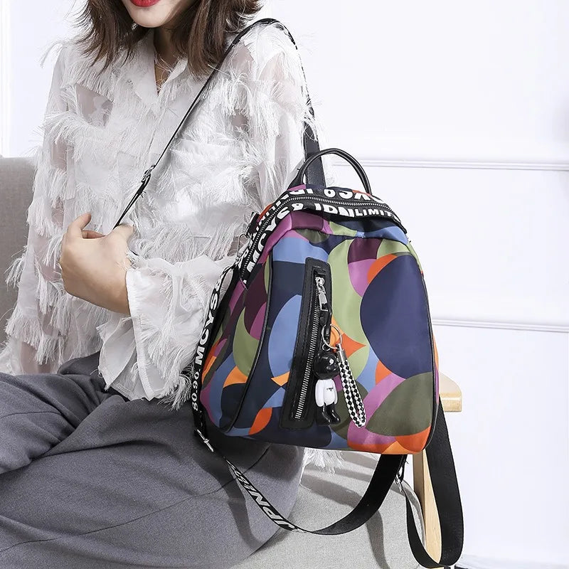 A woman wearing a white sheer blouse and gray pants is holding a purple and green patterned backpack.