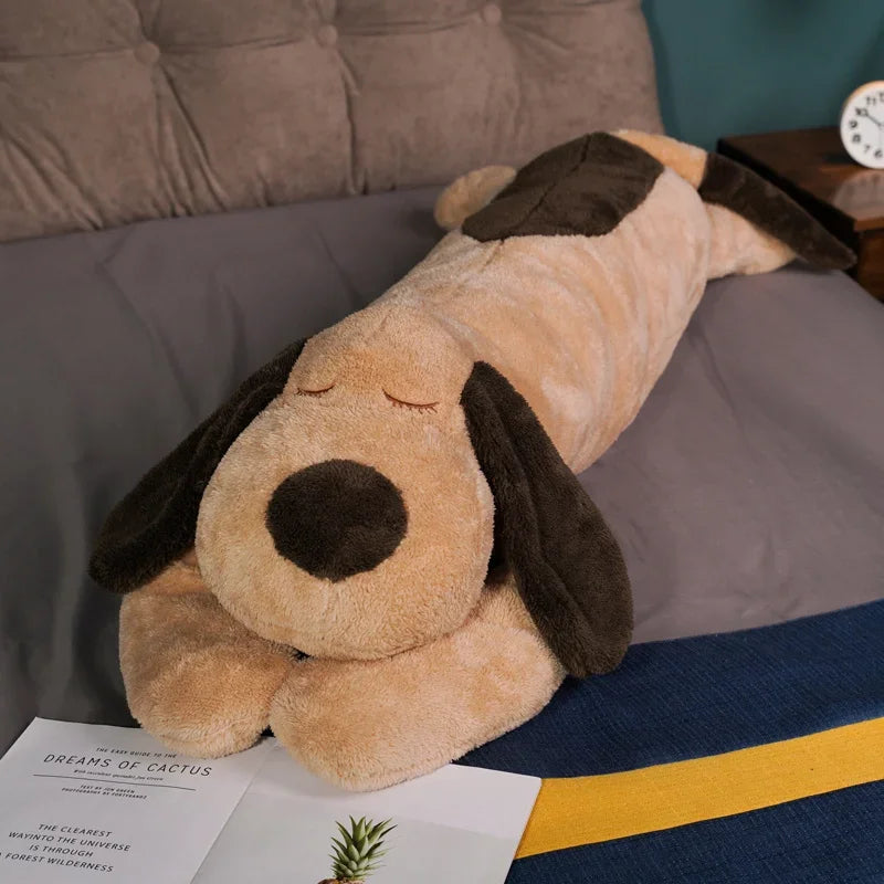 An extra-large soft dog plush pillow with brown and tan fur is resting on a gray bed.