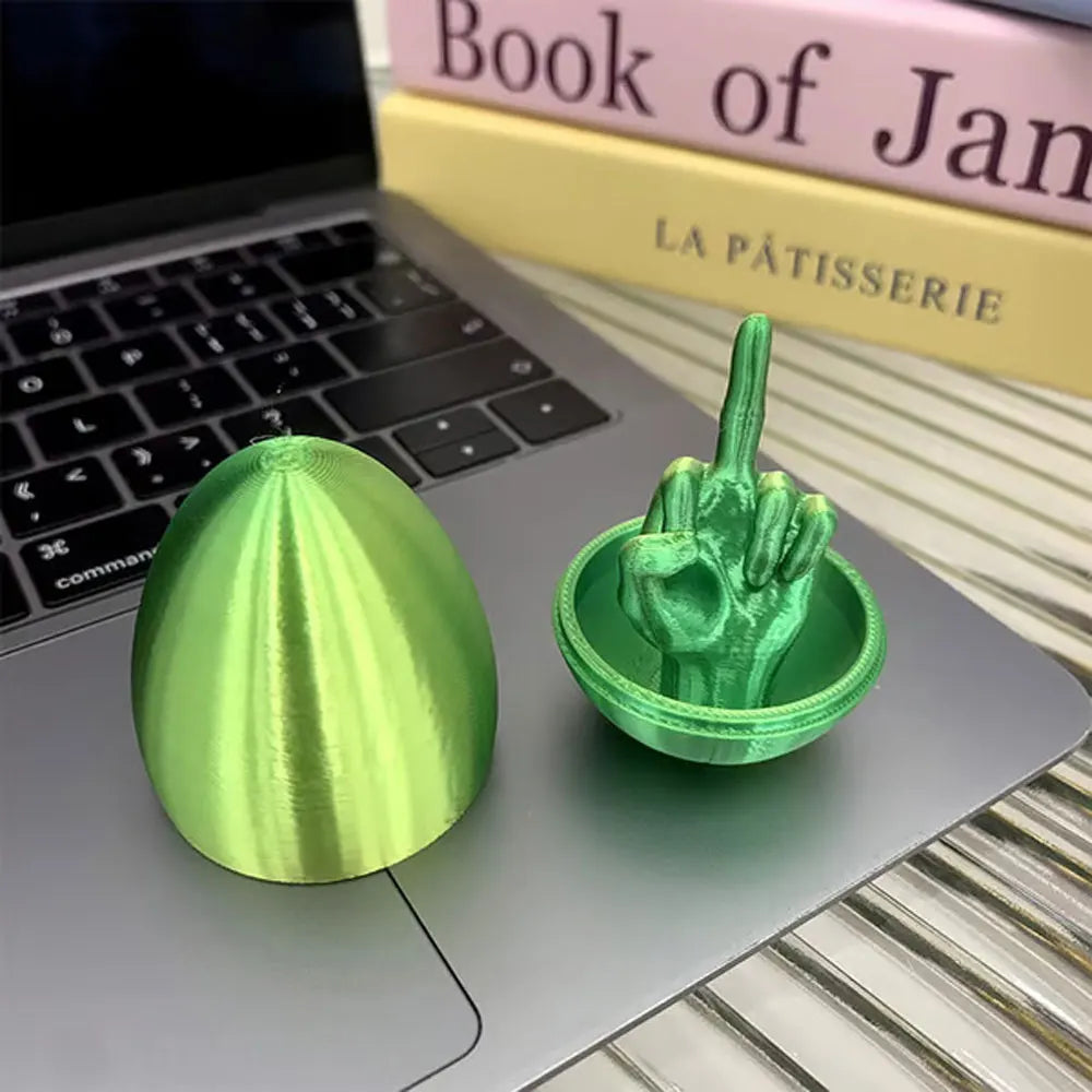 A green Easter egg shaped like a middle finger is placed on a laptop next to a book.