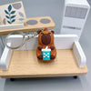 An adorable brown bear keychain charm with a teal ice cube and a green leaf design, resting on a wooden tray.