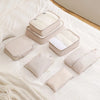 A set of beige and white travel packing cubes and storage organizer bags arranged on a white bed with a knit blanket.