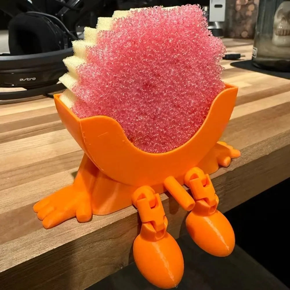 An orange 3D sponge holder with a pink, hanging design is shown on a wooden surface.