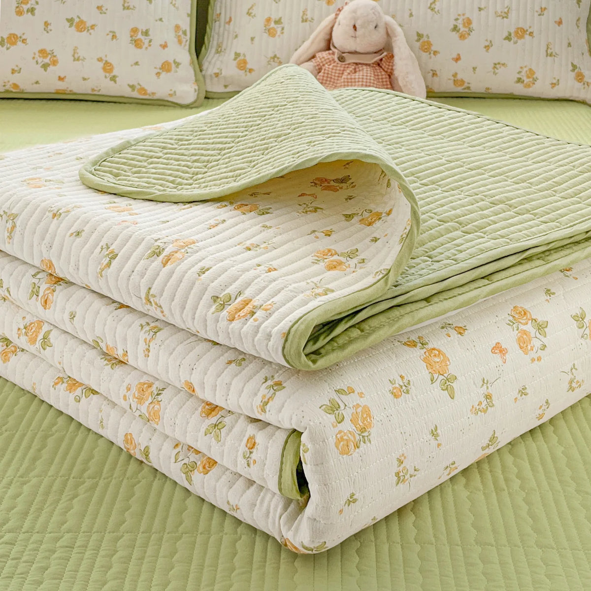 A stack of light green and cream floral quilted linens with a small stuffed rabbit on a pale green background.
