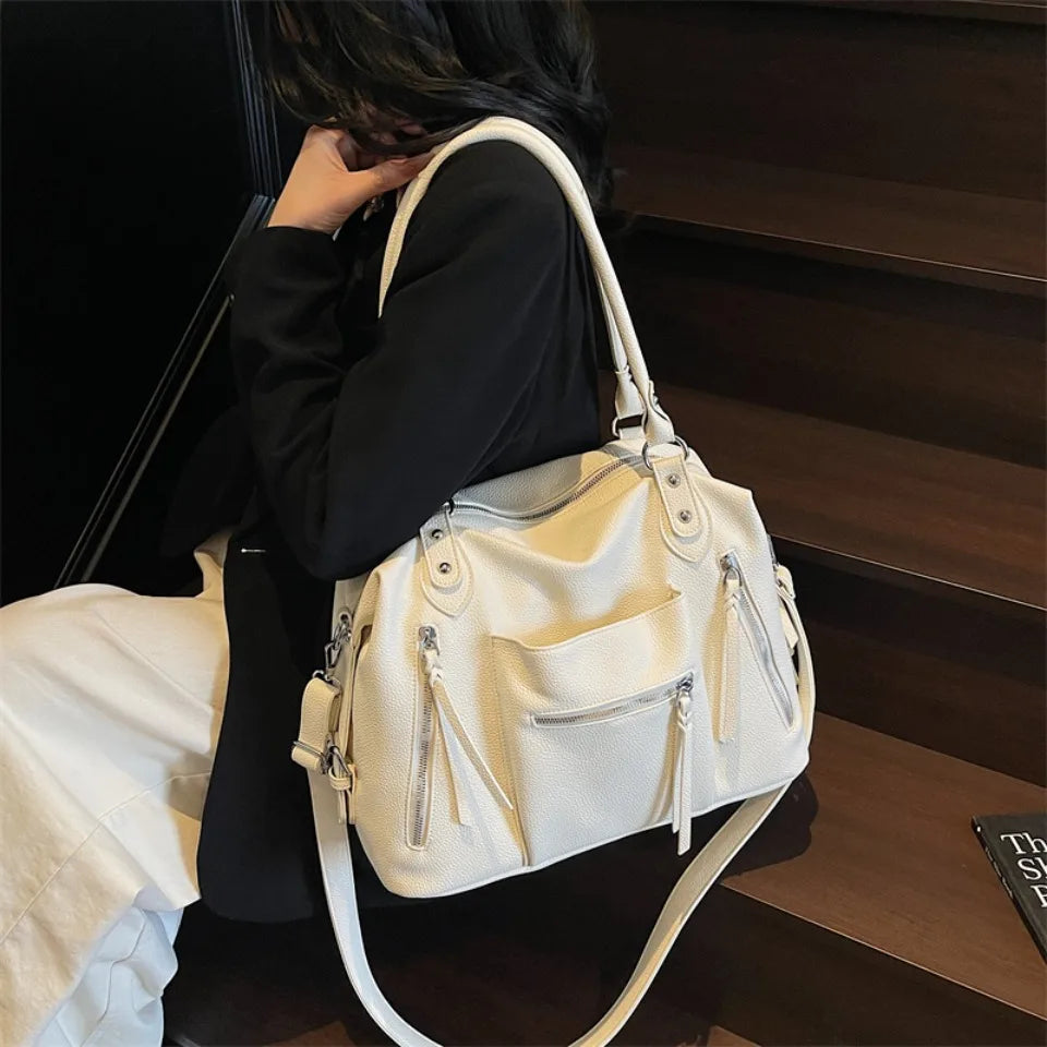 A woman holds a cream-colored soft leather plaid crossbody bag with a white strap against a dark background.