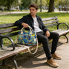 A young man sits on a wooden bench holding an insulated picnic lunch bag with a cross-body strap.