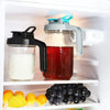 A clear glass jar filled with red liquid is placed next to a white dispenser bottle on a shelf, with lemons and dark grapes n