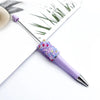 A purple ballpoint pen with a sparkly diamond sequin top rests on a white background next to a piece of paper featuring a gre