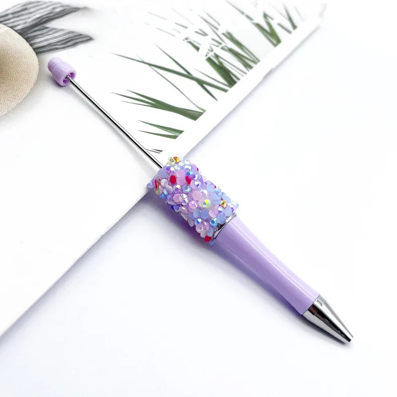 A purple ballpoint pen with a sparkly diamond sequin top rests on a white background next to a piece of paper featuring a gre