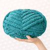 A hand is holding a teal chunky chenille yarn ball.