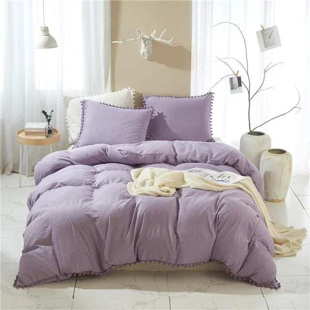 A purple microfiber duvet cover set with pom tassels displayed on a bed against a light wall.