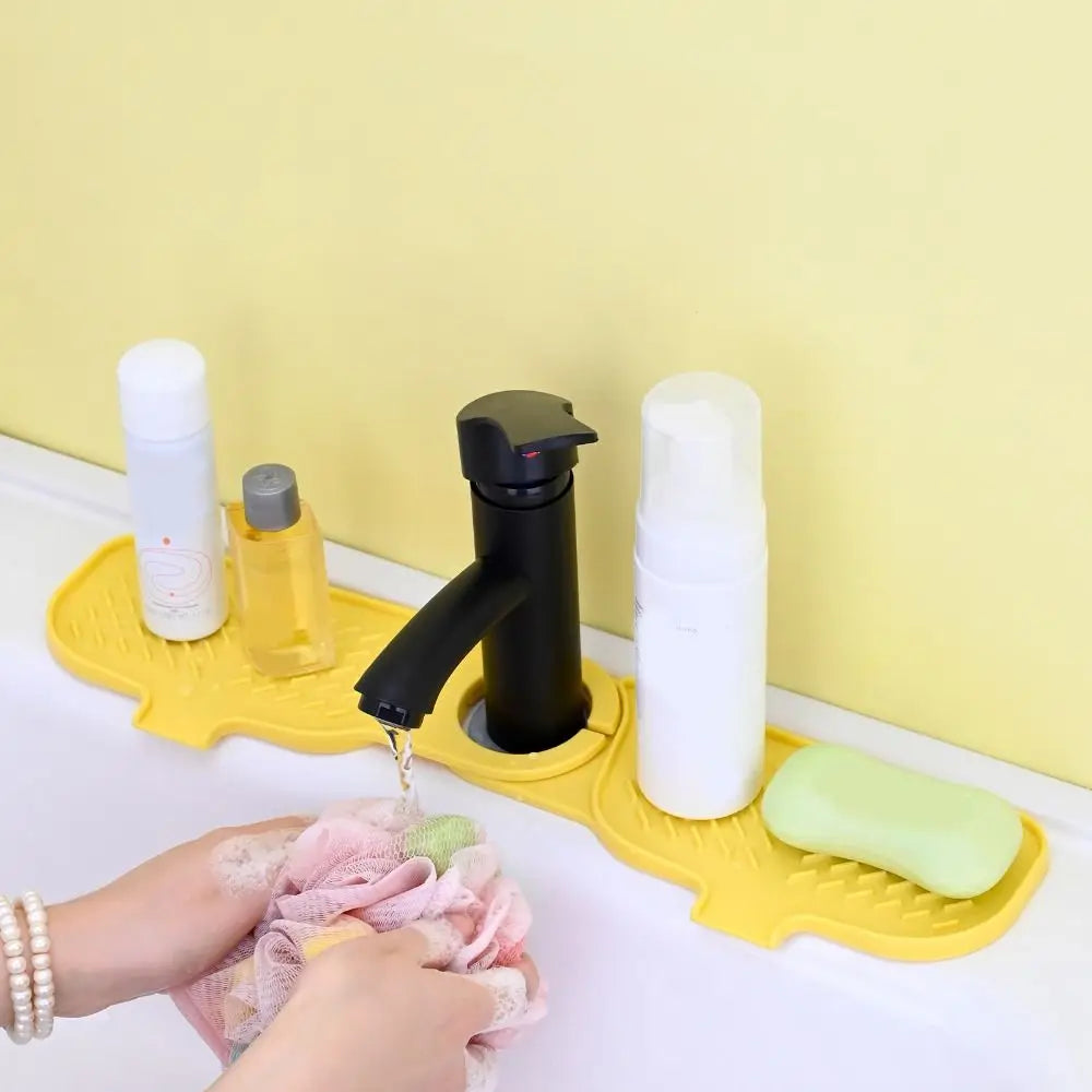 A hand is shown placing a pink cloth onto a yellow silicone sink faucet mat with a black faucet and white soap dispensers.