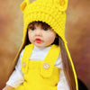 A yellow knitted fox hat baby doll with long brown hair wearing a white shirt and yellow overalls.