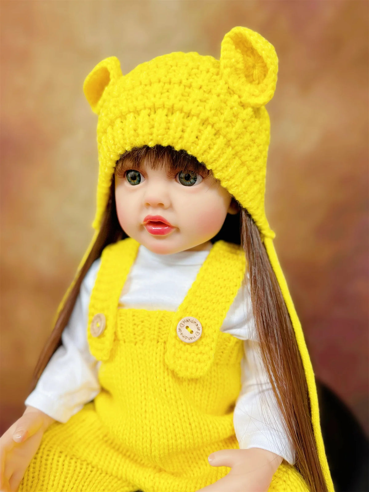 A yellow knitted fox hat baby doll with long brown hair wearing a white shirt and yellow overalls.