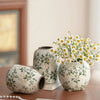 Weiße Keramikvase mit grünen Blumenmustern auf Holzoberfläche neben einer Perlenkette.