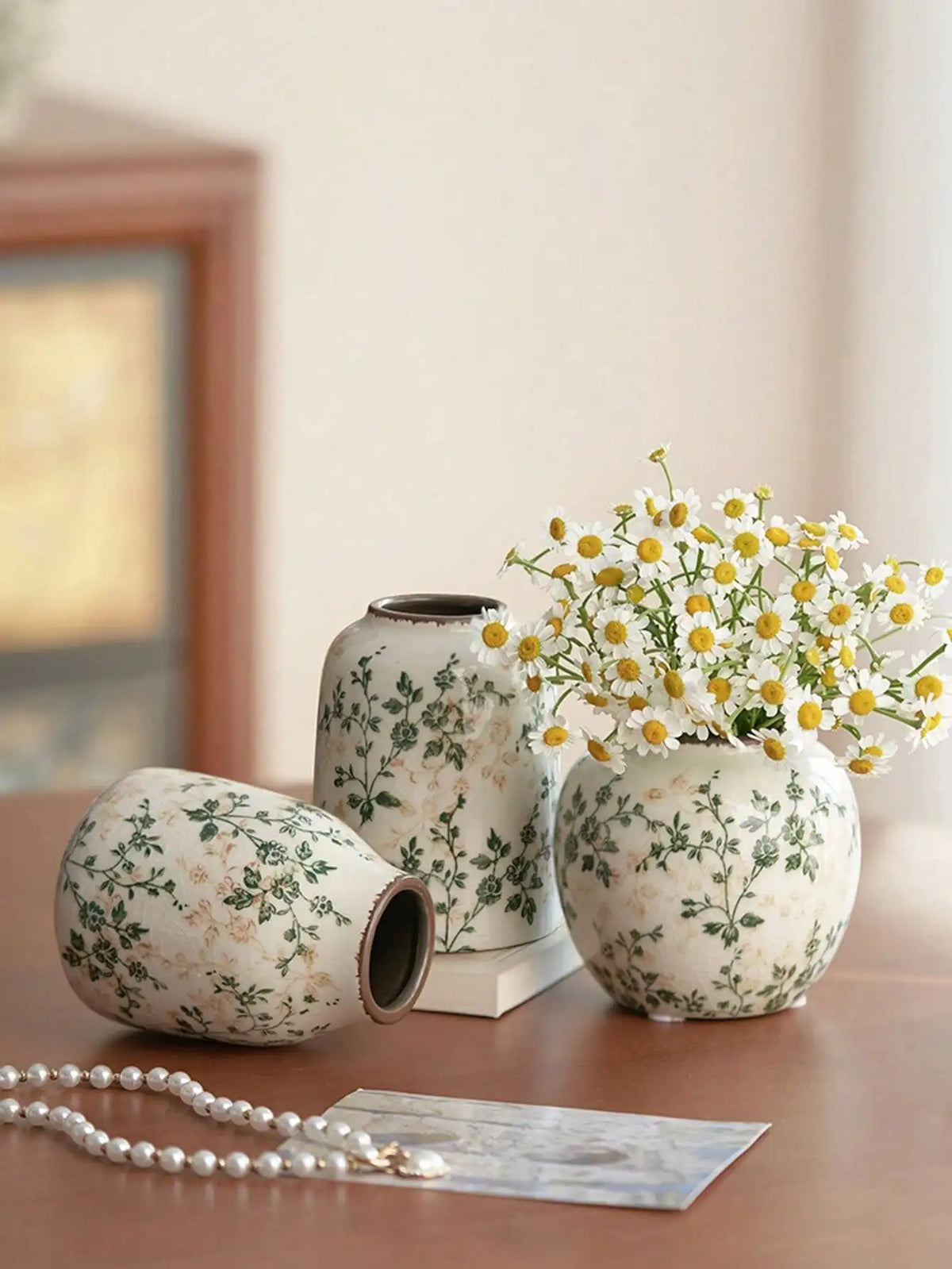 Weiße Keramikvase mit grünen Blumenmustern auf Holzoberfläche neben einer Perlenkette.
