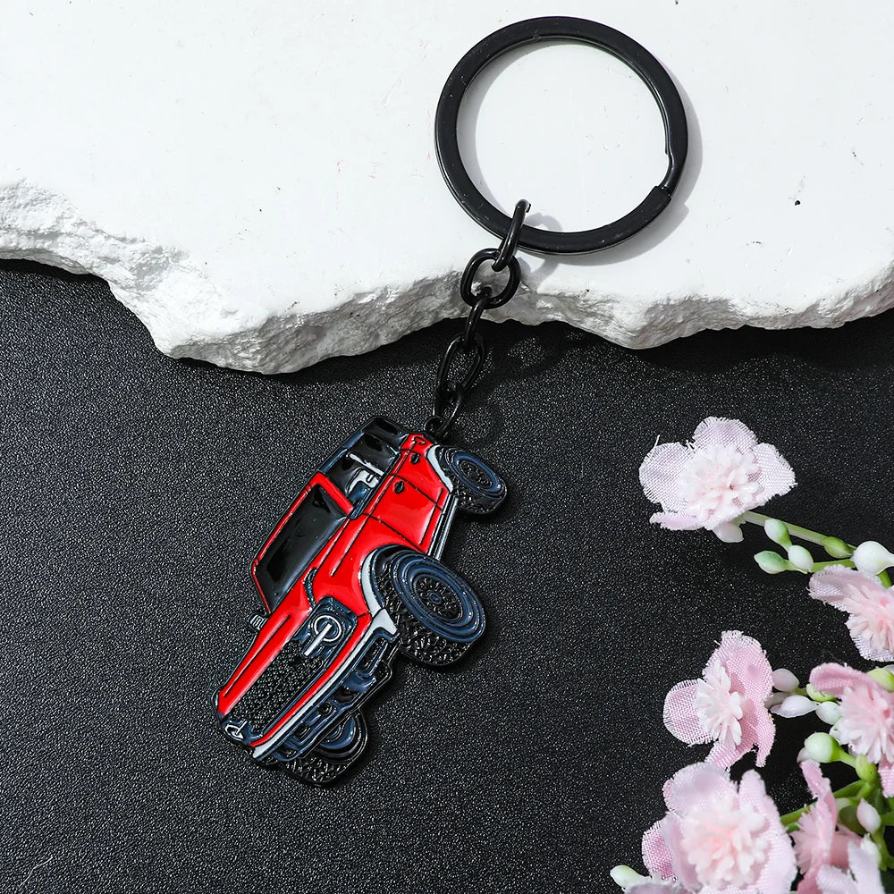A red Ford Bronco zinc alloy keychain attached to a black ring, resting on a dark surface next to pink flowers.