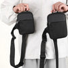 A black canvas crossbody sling bag being held by a person against a white shirt.