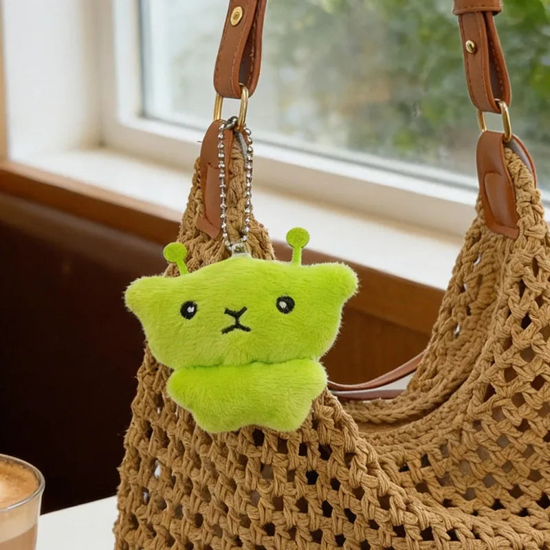 A bright lime green alien plush charm with antennae is attached to a brown woven bag strap.