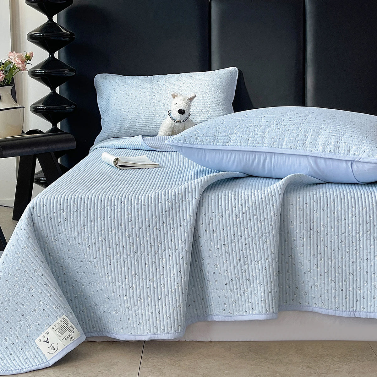 Light blue quilted bedspread with a textured pattern draped over a bed, next to a small white dog.