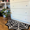 A black and white decorative rug featuring a spider web design with a ghost motif placed on a wooden floor.