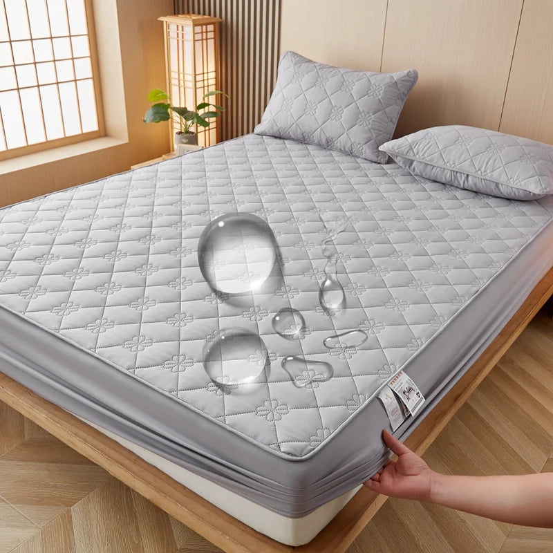 A gray quilted mattress protector is displayed on a wooden bed frame with various glass-like objects on the surface.
