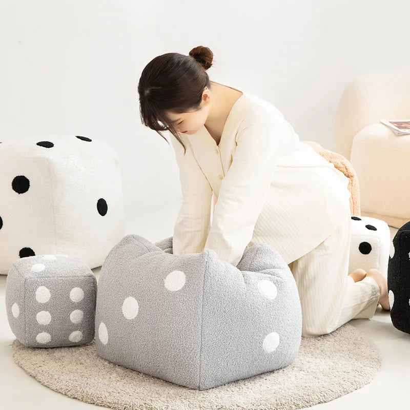 A woman is sitting on a beige rug next to a large gray dice plush pillow and a white polka-dotted cushion.