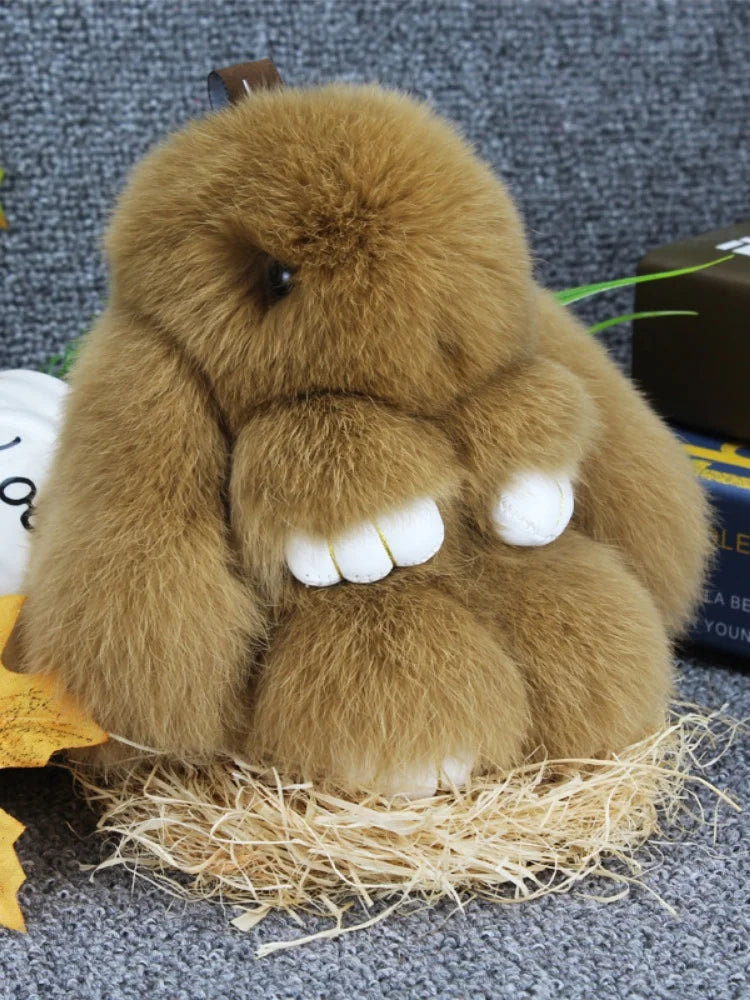 A fluffy brown bunny keychain with a pom pom tail resting on straw.