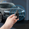 A hand holds a slim carbon fiber car key cover near the headlight of a dark gray car.
