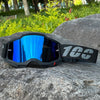 A pair of black motocross goggles with blue mirrored lenses and a black strap resting on a gray rock.
