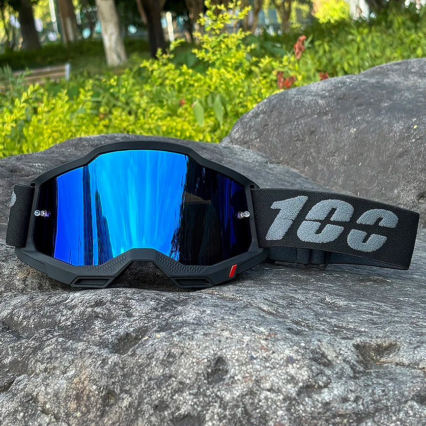A pair of black motocross goggles with blue mirrored lenses and a black strap resting on a gray rock.