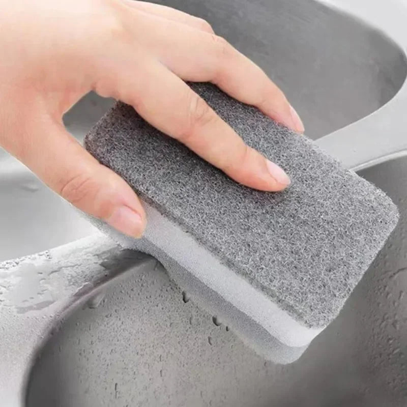 A hand is holding a gray, reusable double-sided cleaning sponge inside a gray sink.