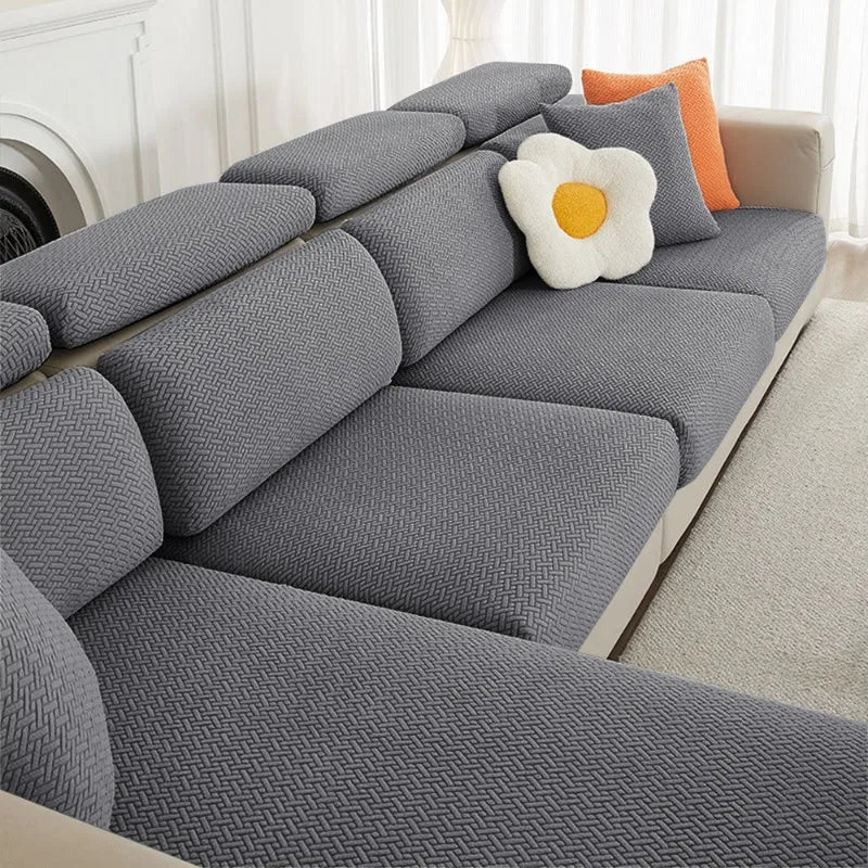 A gray jacquard elastic sofa slipcover with a white flower cushion and an orange accent pillow.