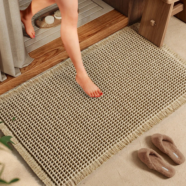 A beige waffle weave bath mat with tassels is placed on a wooden floor next to a door.
