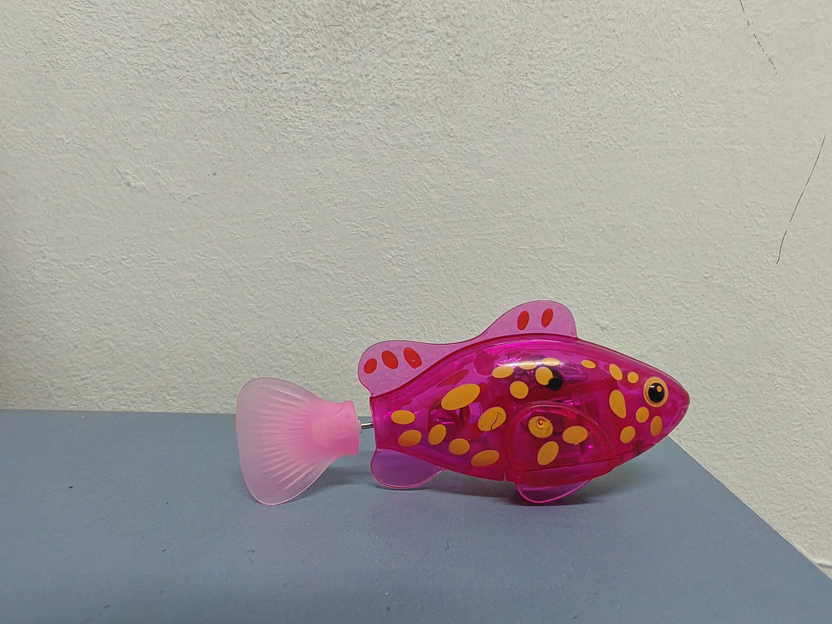 A pink light-up sensor swimming fish toy with yellow spots resting on a gray surface.
