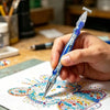 A hand holds a blue diamond painting pen with a silver tip, drawing a colorful heart design on white paper.