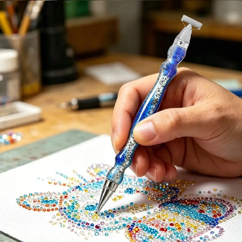 A hand holds a blue diamond painting pen with a silver tip, drawing a colorful heart design on white paper.