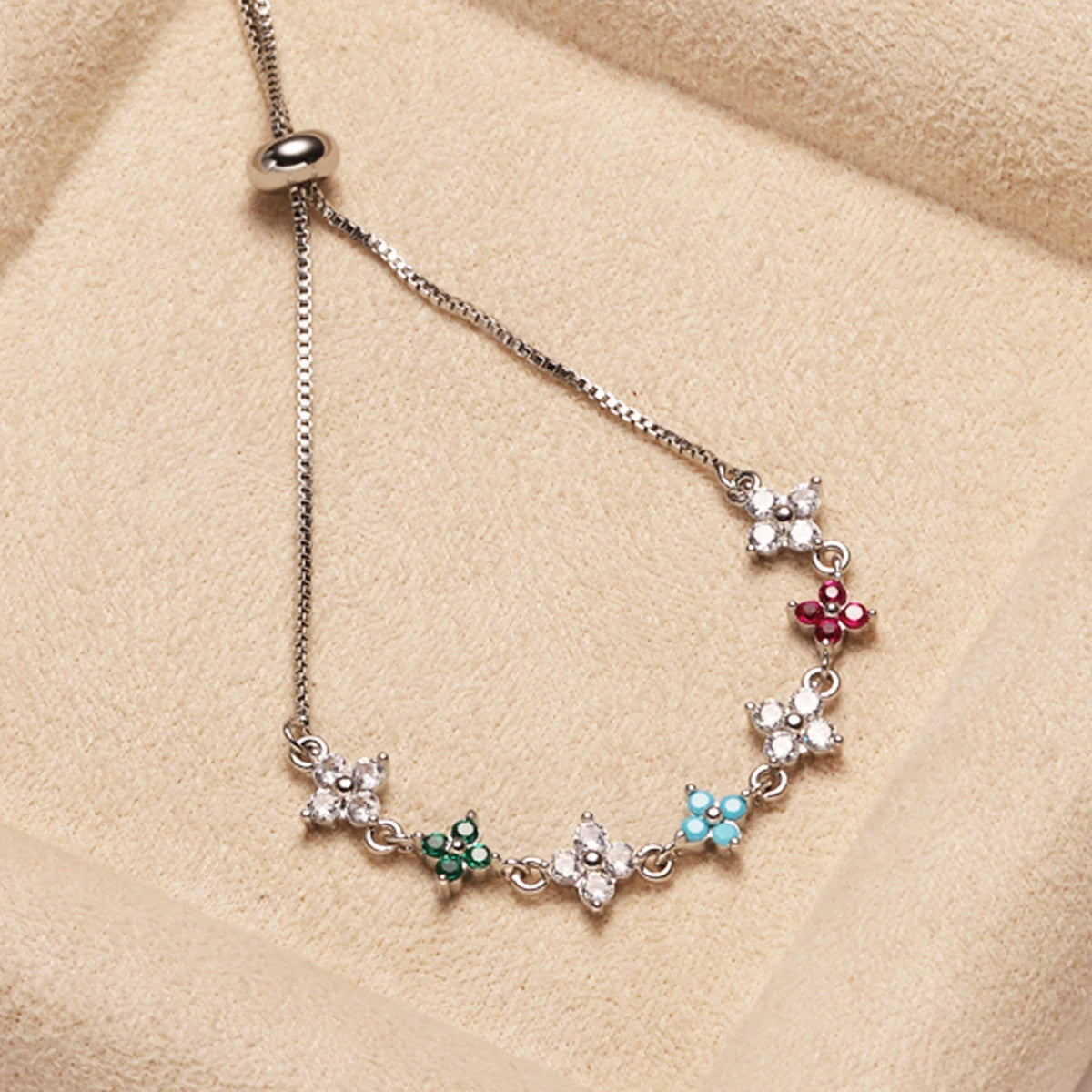 A delicate silver chain bracelet featuring small floral charms and cubic zirconia stones on a beige background.