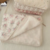 A soft floral muslin baby blanket in cream and pink patterns is shown folded on a light fabric background.