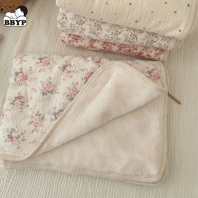 A soft floral muslin baby blanket in cream and pink patterns is shown folded on a light fabric background.