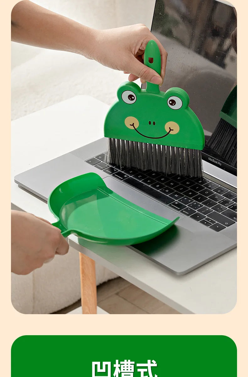 A hand is using a green cartoon broom and dustpan set to clean a laptop or desk surface.