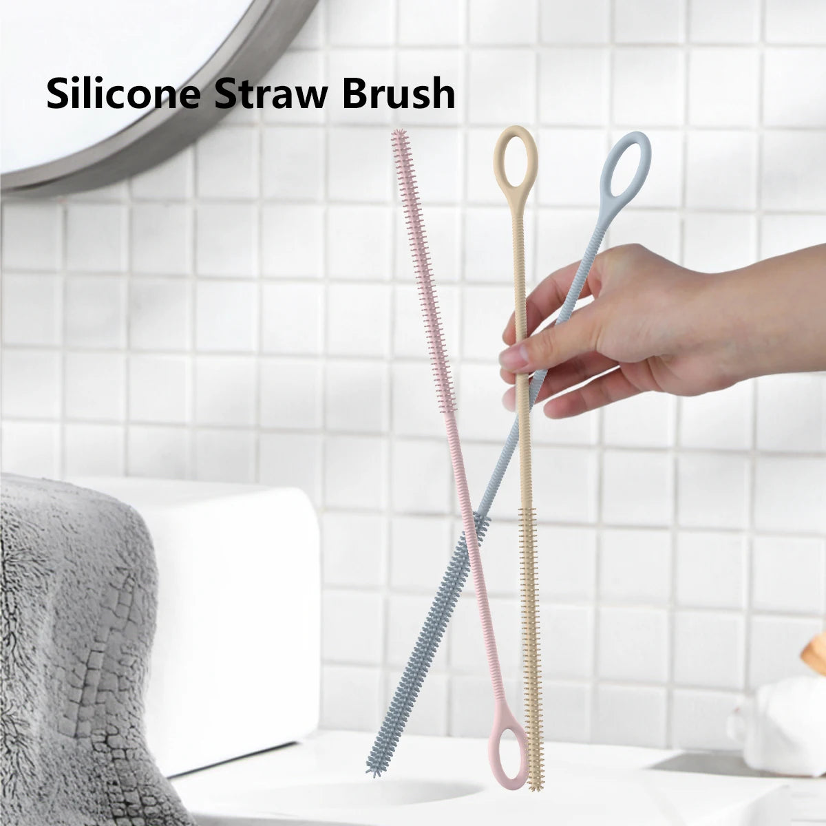 A hand holds a set of two silicone straw cleaning brushes, one pink and one light blue, against a white tiled background.