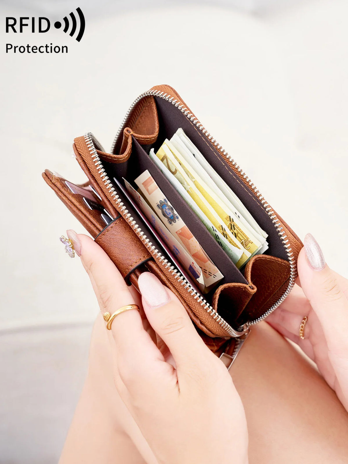 A hand holds a brown vintage RFID blocking short wallet with a zipper closure, showing several cards inside.