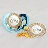 A custom name pacifier featuring a white center and gold rhinestone border, attached to a blue clip.
