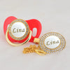 Gold and rhinestone pacifier ring with a white disc engraved with the name Lina set in a red clip.